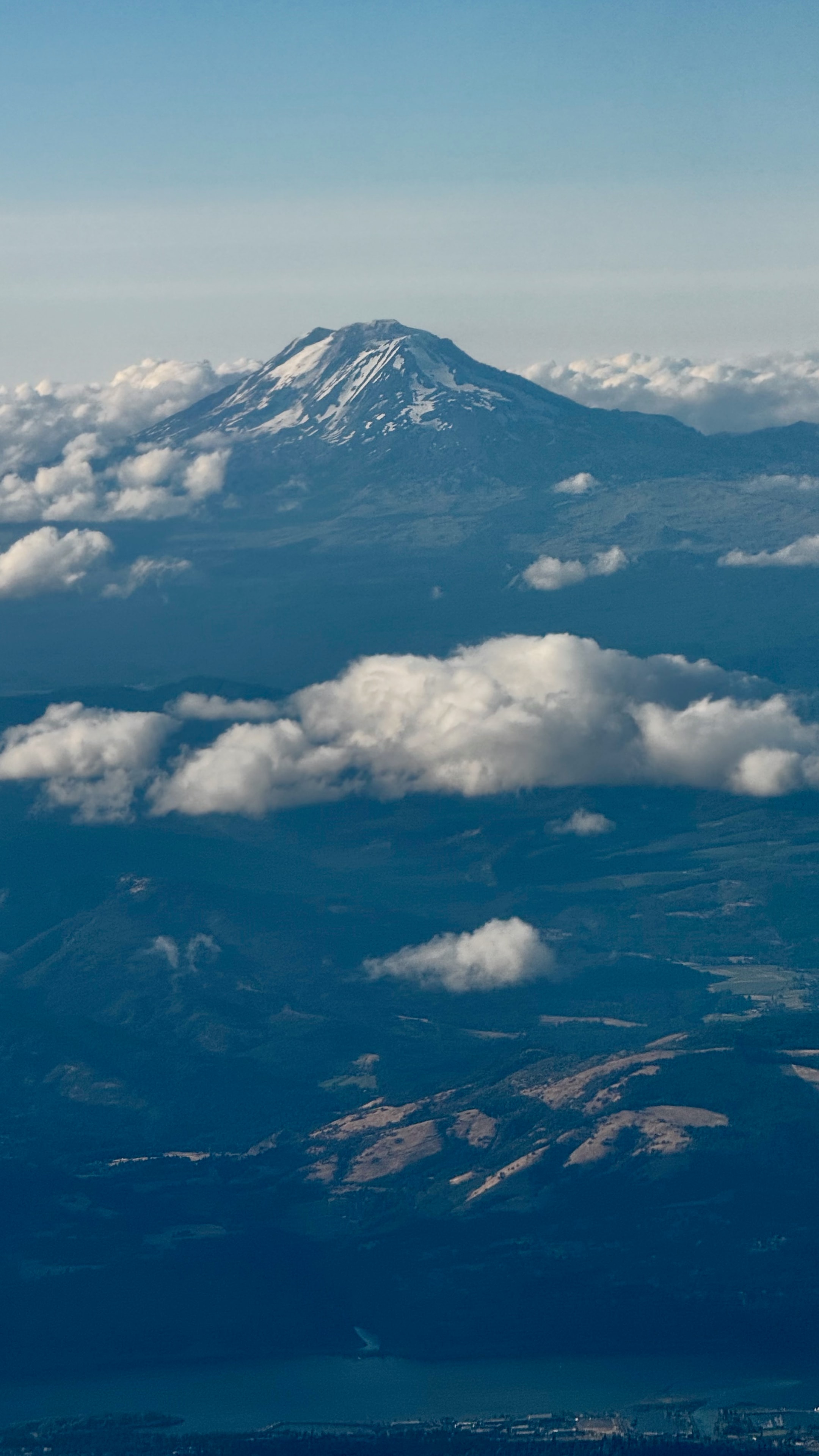 Snow-capped volcanic mountain rising above scattered clouds with green valleys below, aerial view