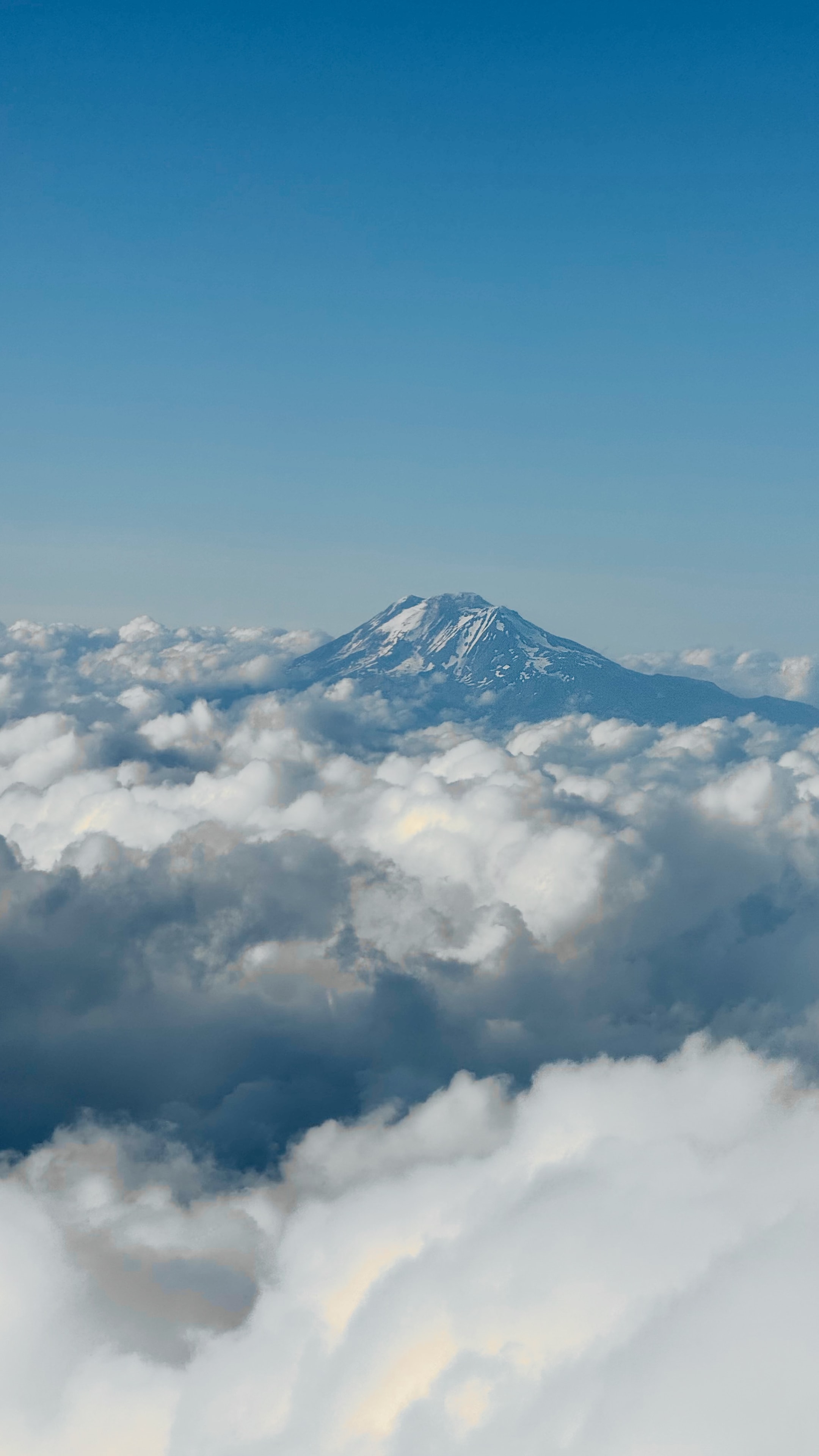 Snow-capped mountain peak emerging from a billowing sea of clouds under clear blue sky, aerial view