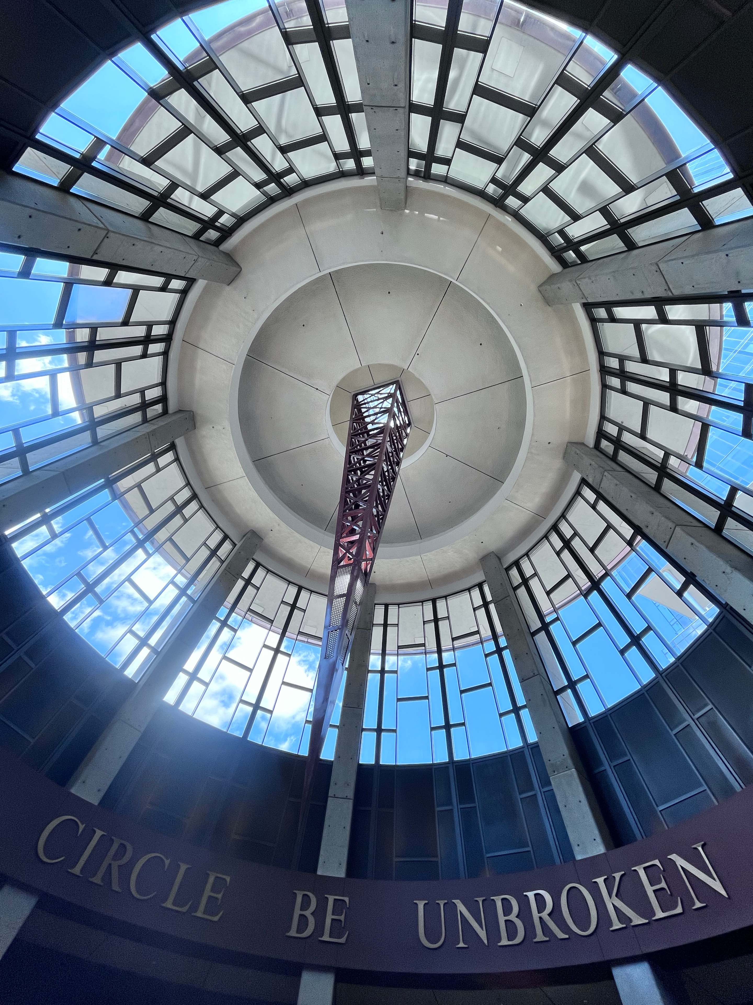 Circular stairwell interior with "Circle Be Unbroken" text and blue sky through glass ceiling